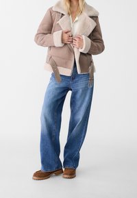 Bruine shearling-voering jas met een tan buitenlaag, voorzien van een brede kraag, ritssluiting en verstelbare riem. Gecombineerd met losse blauwe jeans en bruine laarzen.
