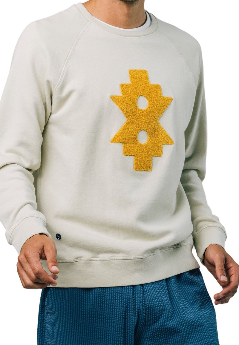 Man in een off-white sweatshirt met lange mouwen, voorzien van een gestructureerd geel geometrisch ontwerp op de borst, en met blauwe gestructureerde broek.
