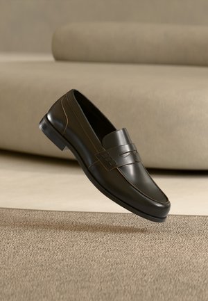 Mustast nahast loafer peene disaini ja dekoratiivse rihmaga, esitatud tekstuurse beeži vaiba ja neutraalse diivani taustal.