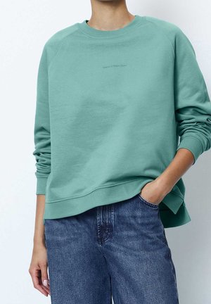 Osoba nosząca morski sweter z okrągłym dekoltem z haftowanym napisem „Marc O’Polo Denim” oraz niebieskie jeansy, stojąca z jedną ręką w kieszeni.