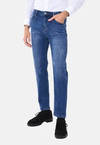 Jeans in denim blu con vestibilità dritta, lievi segni di usura, cinque tasche e chiusura con bottone. Indossati con scarpe in suede nere.