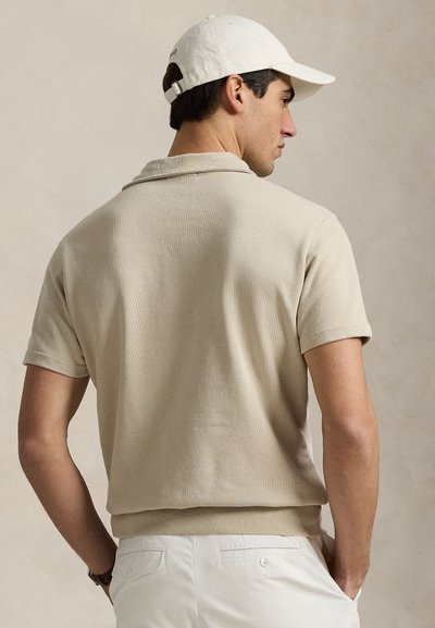 Mand iført beige kortærmet polo, hvide bukser og hvid kasket, stående sidevendt med hænderne i lommerne mod en neutral baggrund.
