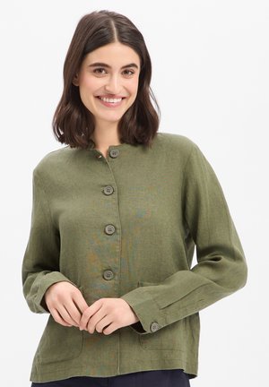 Młoda kobieta uśmiechająca się, ubrana w oliwkowo-zieloną kurtkę zapinaną na guziki z długimi rękawami i przednimi kieszeniami, stoi na białym tle.