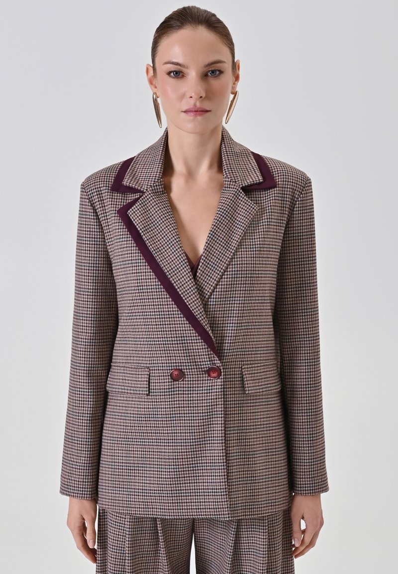 Blazer double boutonnage en motif à carreaux texturé marron et crème, avec des revers et des boutons bordeaux, doté de deux poches avant.