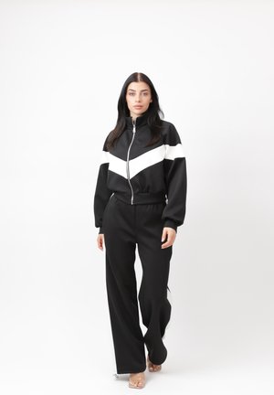 Kvinde går fremad iført en sort og hvid zip-up løbejakke og sorte, vidde bukser mod en ens hvid baggrund.