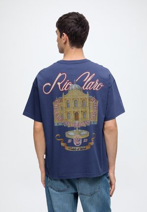 Homme portant un t-shirt bleu marine avec une impression détaillée d'un bâtiment jaune et le texte "Rio Claro" dans le dos, associé à un jean bleu.