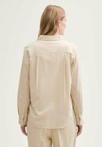Camicia beige a maniche lunghe con colletto, polsini con bottone e taglio dritto, realizzata in un tessuto testurizzato. Cuciture sul retro.