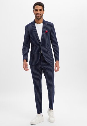 Lachende man die een marineblauw gestreept pak draagt met een rood pochet, wit T-shirt en witte sneakers, staand tegen een effen achtergrond.