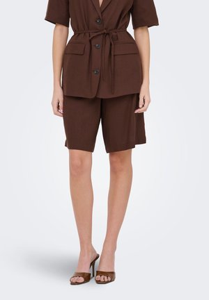 Kvinde iført brun knappet blazer med bælte, matchende knælange shorts og brune højhælede åbne sandaler.