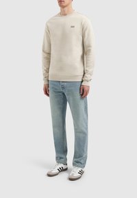 Beige crewneck sweatshirt met geribbelde manchetten en zoom, voorzien van geborduurde tekst "PURE PATH" op de borst, gecombineerd met lichtblauwe spijkerbroeken en sneakers.