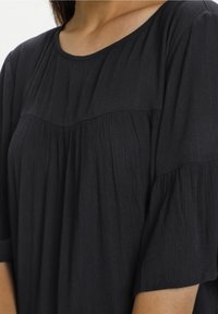 Mujer vestida con una blusa negra texturizada de mangas cortas con detalles plisados y un escote redondeado, mostrada de cuello a medio torso.