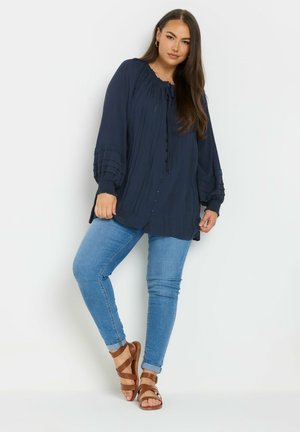 Blouse bleu marine à manches longues avec texture plissée et fermeture à boutons, associée à un jean bleu clair et des sandales marron.