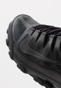 Schwarzer Sportschuh mit Mesh-Obermaterial, strukturiertem Muster, runder Zehenpartie und Gummisohle mit kreisförmigem Profil für Grip.