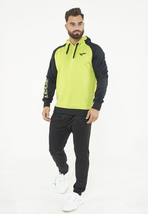 Uomo in piedi che indossa una felpa con cappuccio giallo neon e nero, pantaloni neri e sneakers bianche su sfondo bianco.