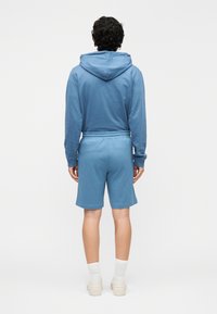 Blå huvtröja med dragsko och långa ärmar, kombinerad med matchande shorts. Mjukt tyg, ledig passform, bärs med vita ankelstrumpor och sneakers.