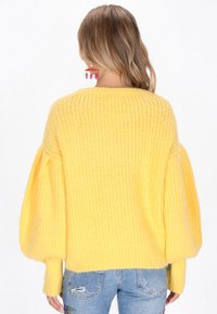 Gelbes Strickpullover mit lockerer Passform, Ballonärmeln und strukturiertem Stoff. Kombiniert mit hellblauen, ausgefransten Jeans, die von hinten gezeigt werden.