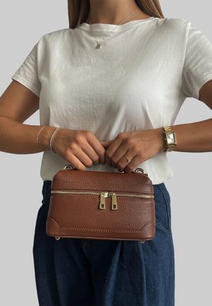 Una borsa a tracolla in pelle marrone con cerniera e accessori dorati. Forma rettangolare con finitura testurizzata, tenuta da una persona che indossa una camicia bianca.
