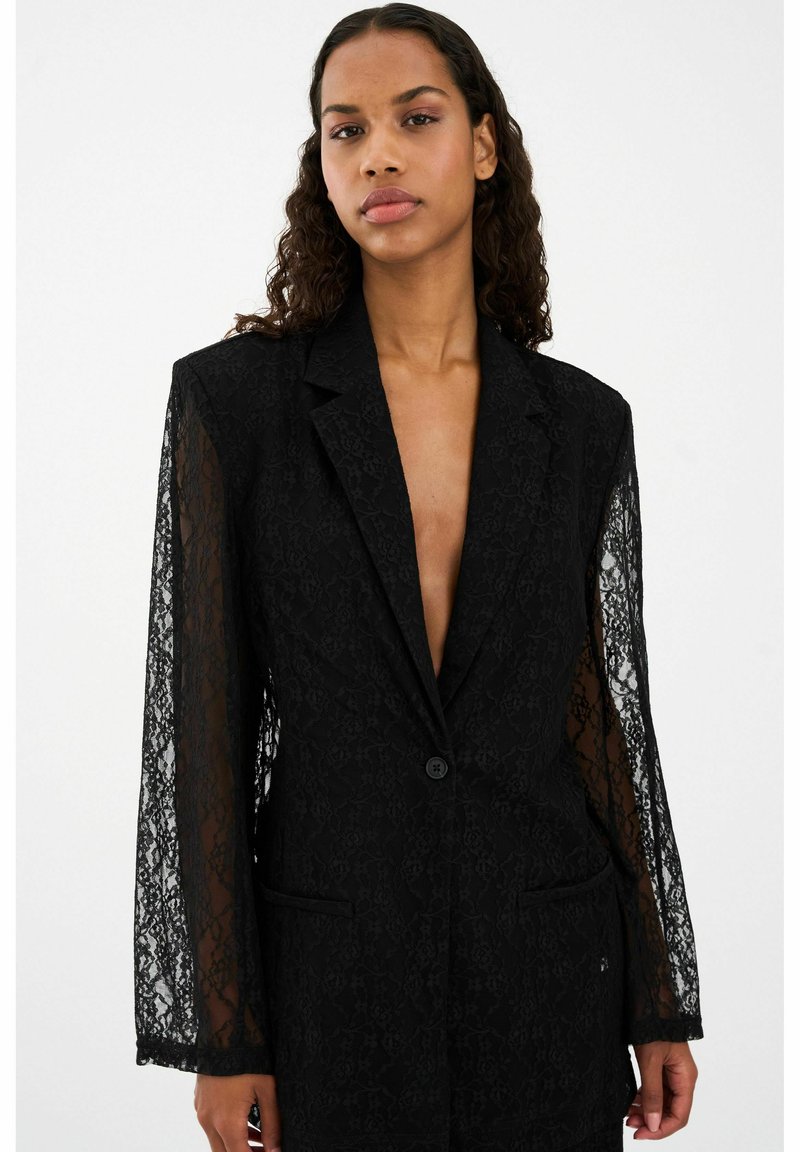 Must mustriga bleiser, millel on pikad läbipaistvad varrukad, ühe nupuga kinniti, tähni krae, kaks esist nii taskut ja keerukad lillemustrid.