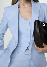 Helles blaues Leinenanzug mit tailliertem Blazer und Weste, der über zwei Knöpfe verfügt. Hält eine schwarze Handtasche aus Leder mit geschwungenen Kanten.