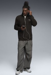 Felpa nera con zip a metà, pantaloni cargo grigi e sneakers bianche e nere. L'individuo utilizza uno smartphone e cuffie.