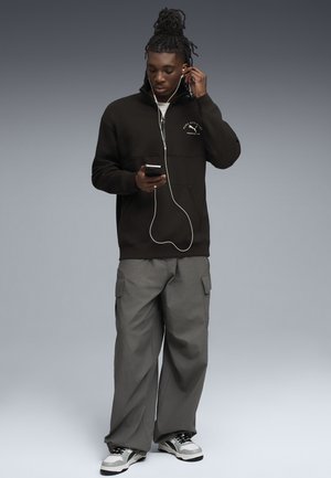 Svart hoodies med halv dragkedja, grå cargobyxor och svartvita sneakers. Personen använder en smartphone och hörlurar.