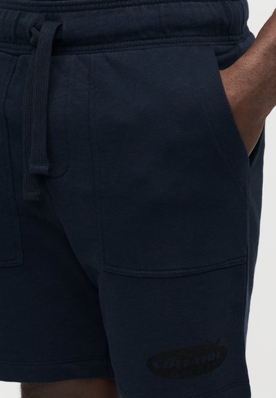 Σκούρα μπλε βαμβακερή βερμούδα με ελαστική ζώνη μέσης, κορδόνι, πλαϊνές τσέπες και διακριτική γραφική λεπτομέρεια στο πόδι. Ομαλή υφή.