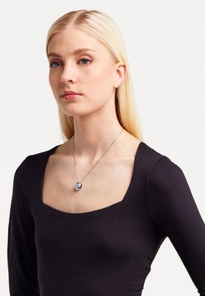 Zilveren ketting met een ronde hanger, voorzien van een glanzend zwart middenstuk, gedragen door een model met lang, steil blond haar, gekleed in een zwarte top.
