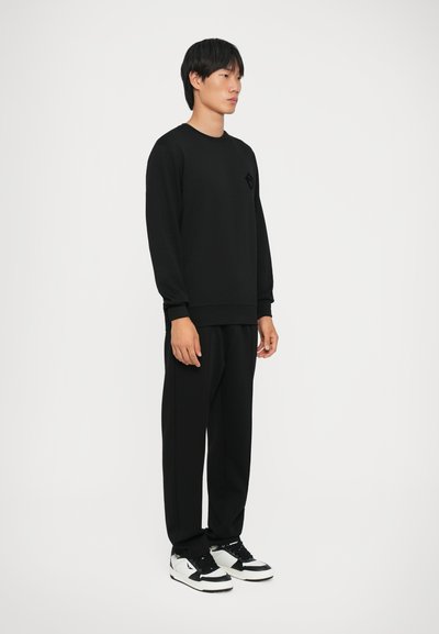 Felpa nera con collo rotondo, polsini a coste e una leggera texture. Abbinata a pantaloni neri larghi e sneakers bianche con dettagli neri.