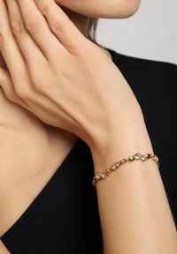 Pulsera de oro que presenta una mezcla de cuentas de oro redondas y piedras de strass claras, colocada sobre una muñeca desnuda. Llevada con una blusa negra.