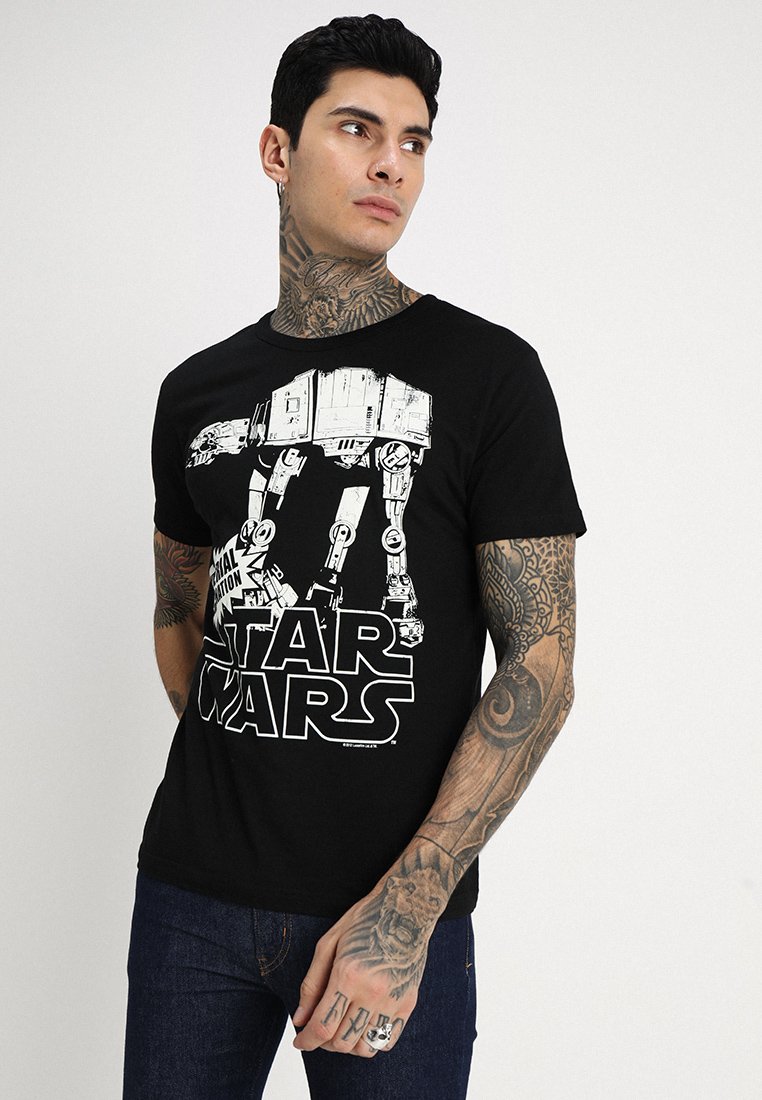 Mann mit Tätowierungen, der ein schwarzes Star Wars T-Shirt mit einem großen weißen AT-AT-Walker-Logo trägt, steht vor einem schlichten hellen Hintergrund.