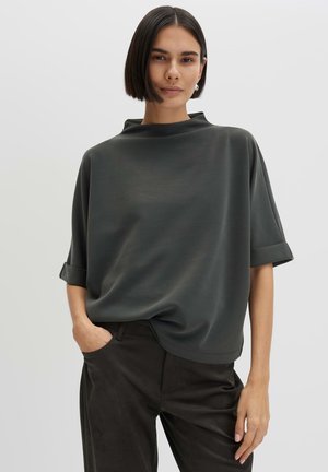 Vrouw met kort, steil donker haar die een losse donkergrijze top met korte mouwen en zwarte broek draagt, met één hand in de zak tegen een effen achtergrond.