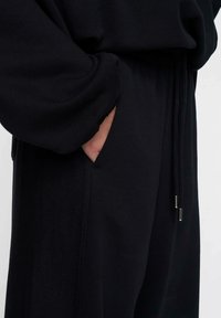 De hand van een persoon bevindt zich gedeeltelijk in de voorkant van de zwarte sweatpants met een trekkoord in de taille en textuurpanelen aan de zijkanten.