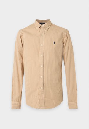 Homme portant une chemise beige rayée à boutons, un pantalon foncé et des mocassins marron foncé, debout sur fond blanc.