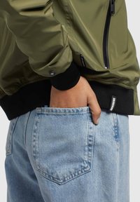 Giacca bomber verde oliva con polsini a costine neri, dotata di una tasca con zip. Jeans in denim con lavaggio blu chiaro e una tasca posteriore singola.