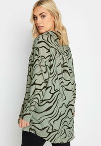 Long-sleeve shirt in light green fabric with abstract black wavy patterns. Features a buttoned cuff and a loose, relaxed fit.