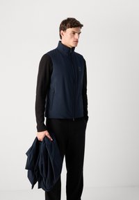 Gilet senza maniche blu navy con colletto alto, chiusura con zip e tasche laterali, realizzato in materiale leggero e morbido, indossato sopra pantaloni neri.
