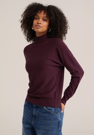 Vrouw met krullend haar die gouden oorringen draagt, een donkerpaarse coltrui en een blauwe spijkerbroek, staand met één hand in de zak.