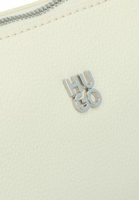 Cuero blanco texturizado con un emblema plateado del logo "Hugo". Detalle de cremallera en la parte superior. El material tiene un acabado suave y graneado.