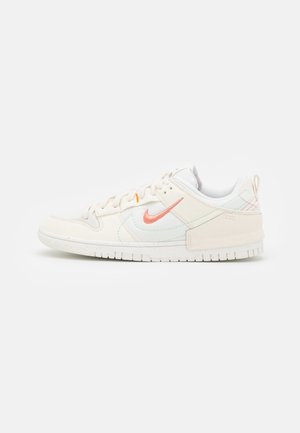 Witte lage Nike sneaker met lichtroze swoosh, vetersluiting en rubberen zool op een effen lichte achtergrond.