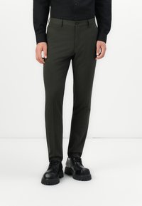 Bărbat îmbrăcat cu pantaloni strânși de culoare verde măslinie, pantofi din piele neagră groși și o cămașă neagră cu mâneci lungi și nasturi, stând pe un podea albă.