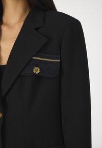 Blazer noir sur mesure avec une poche en tissu brillant dotée d'une fermeture éclair dorée et de détails en bouton, des revers structurés et une texture lisse.