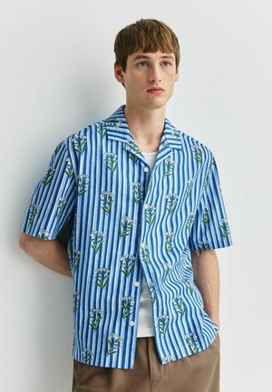 Young man with short brown hair wearing a blue and white striped short-sleeve shirt with white flower patterns, posing against a white wall.