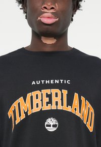 Crna pamučna majica s natpisom "AUTENTIČNI TIMBERLAND" u podebljanom bijelom i narančastom tekstu, s bijelim logotipom drveta sprijeda.