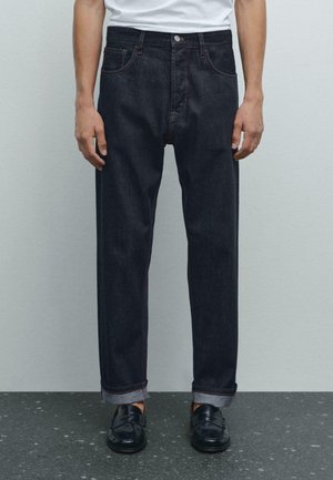 Jeans in denim scuro con taglio dritto, caratterizzati da cuciture arancioni, cinque tasche e orli arrotolati, abbinati a mocassini neri.