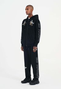 Schwarzer Kapuzenpullover mit Stickerei, der einen Oktopus und Flügel zeigt, kombiniert mit schwarzen Jogginghosen. Beide haben "ED HARDY" aufgedruckt. Schwarze Sneaker.