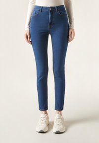Blå denim skinny jeans med hög midja, har fem fickor, beige sömmar och knappstängning. Matchas med vita sneakers.
