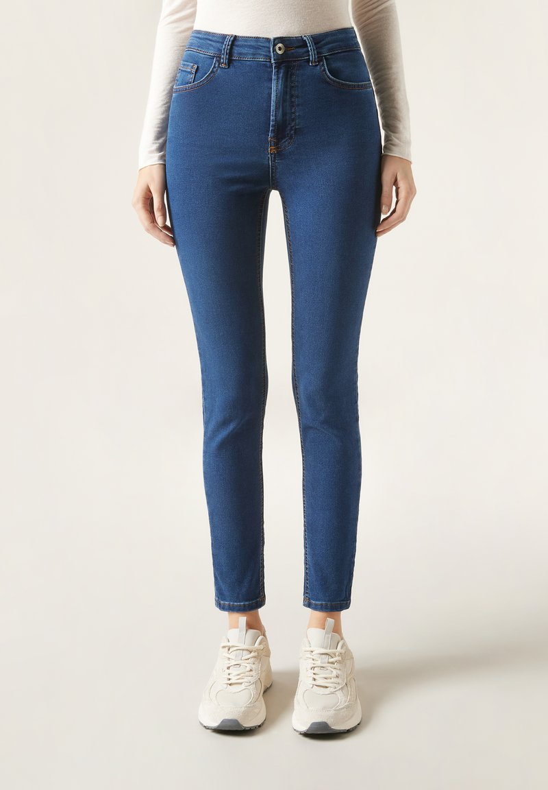 Blå denim skinny jeans med hög midja, har fem fickor, beige sömmar och knappstängning. Matchas med vita sneakers.
