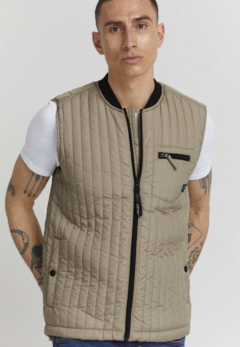 Beige quiltet vest med svart glidelås foran, sidelommer og en liten lomme på venstre bryst. Stoffet har en glatt tekstur og subtil glans.