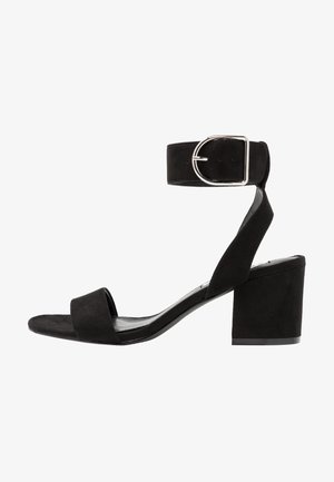 Sandal com salto de camurça preto, com tiras no tornozelo, fivela prateada, bico aberto e salto quadrado. Sola plana e design minimalista.