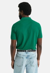 Grünes Poloshirt aus Baumwolle mit klassischem Kragen und kurzen Ärmeln, kombiniert mit hellblauen Jeans und einem gemusterten Gürtel.